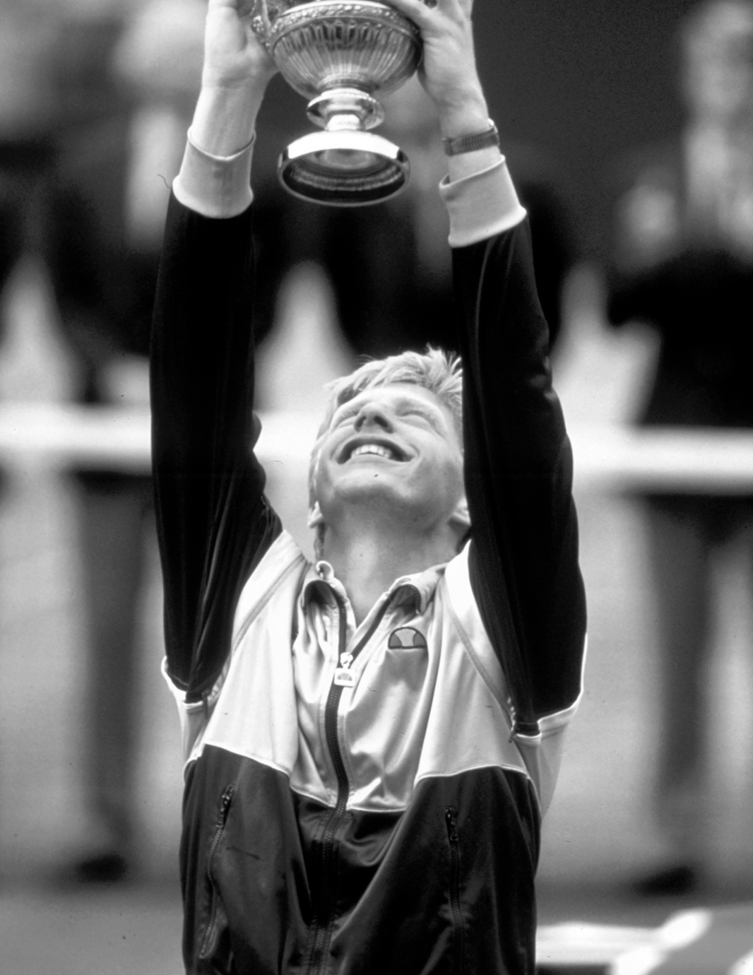 Boris Becker, wearing a Ellesse Track Jacket, holding a trophy above their head in a celebratory pose.